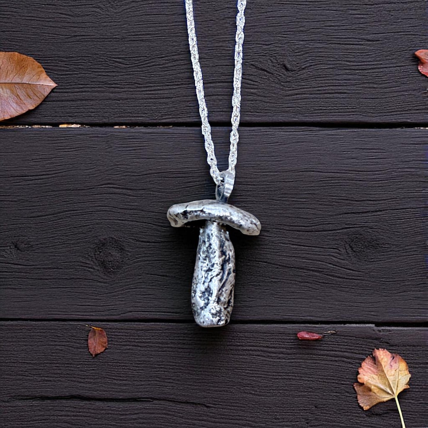 Silver mushroom-shaped pendant necklace on a dark wooden surface with scattered leaves.