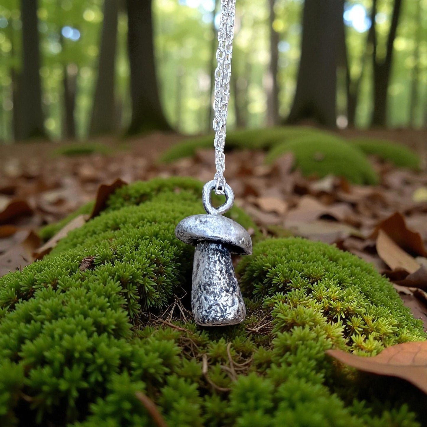 Silver mushroom-shaped pendant on a chain hanging over green moss in a forest setting.
