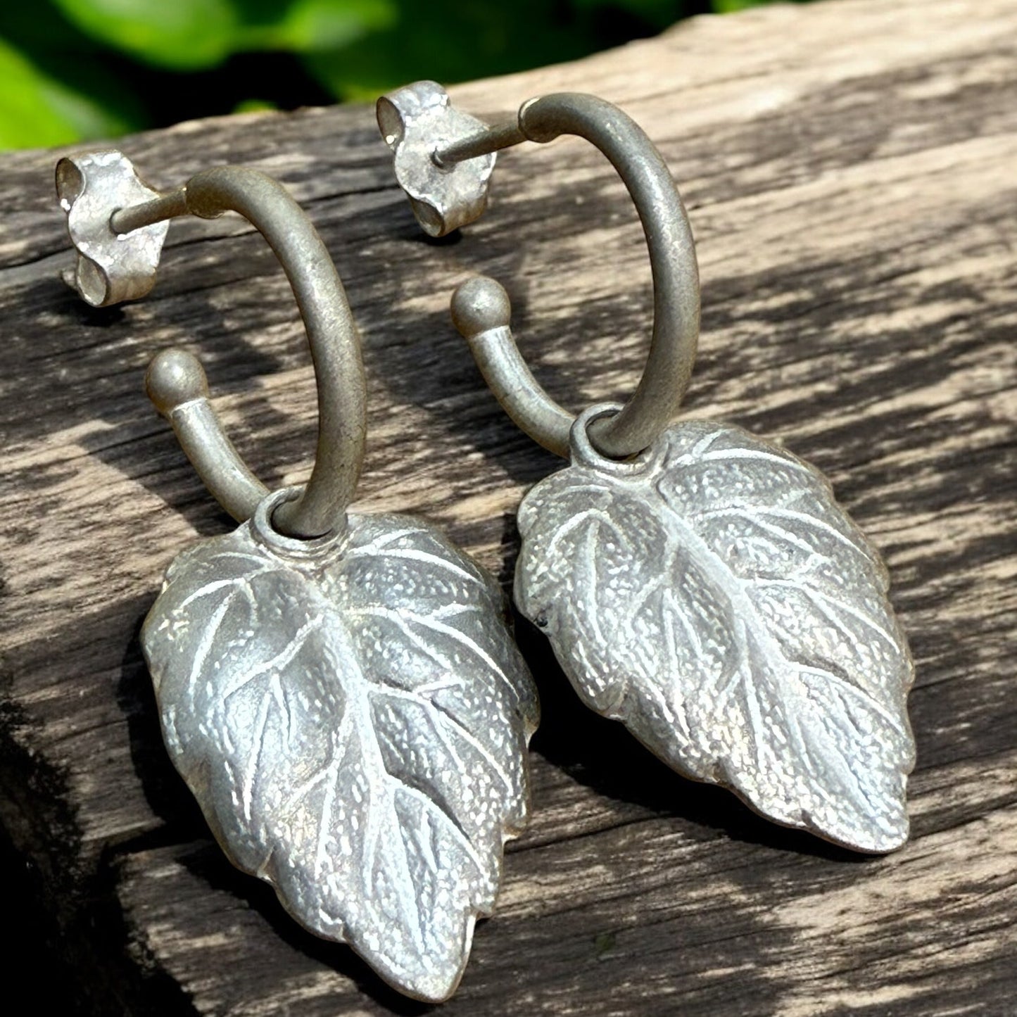 A pair of silver hoope earrings with leaf charms resting on a piece of wood.
