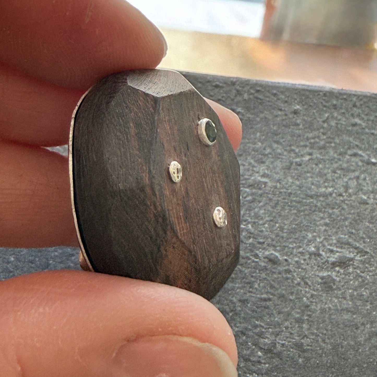 Wooden brooch with sapphire & silver accents held between fingers on a dark surface