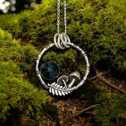 A pendant resting on moss. The pendant is a bamboo style circle with mushrooms, a fern frond and a teal moss kyanite stone