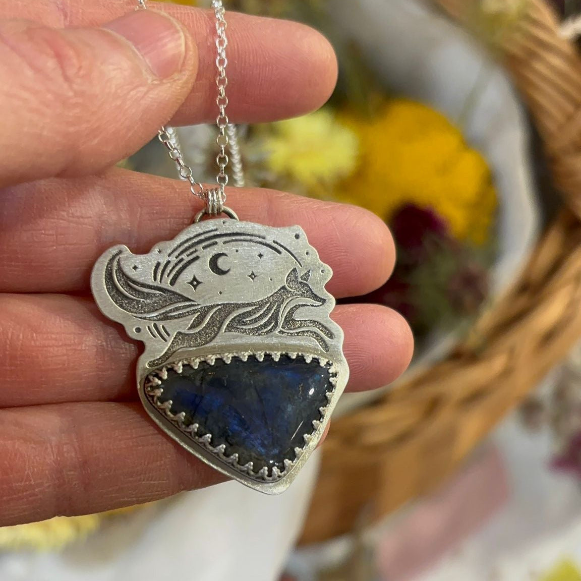 Hand holding a silver necklace with an etched fox pendant against a floral background