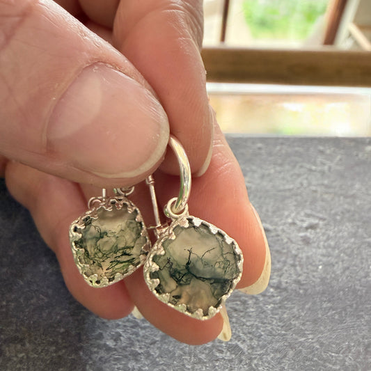 Hand holding a pair of silver earrings with moss agate drops against a neutral background.
