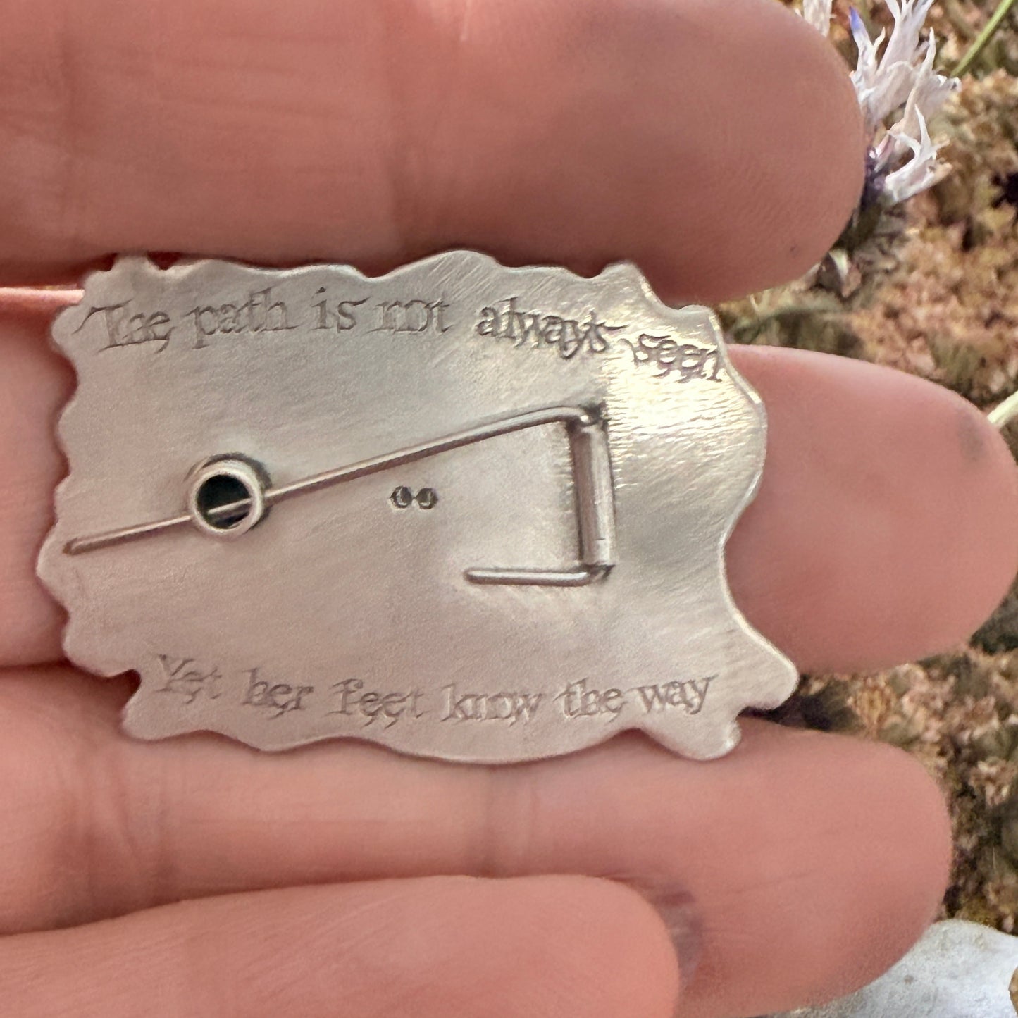 Silver brooch held between fingers with a blurred natural background