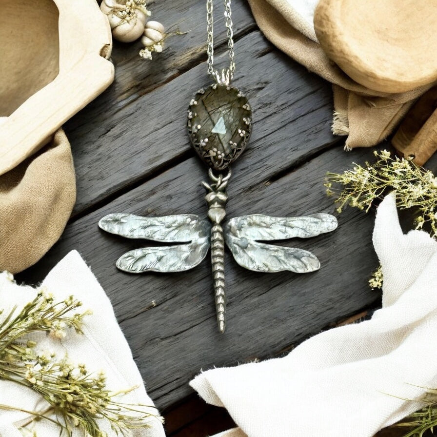 Dragonfly-shaped pendant necklace on a wooden surface with fabric and flowers.