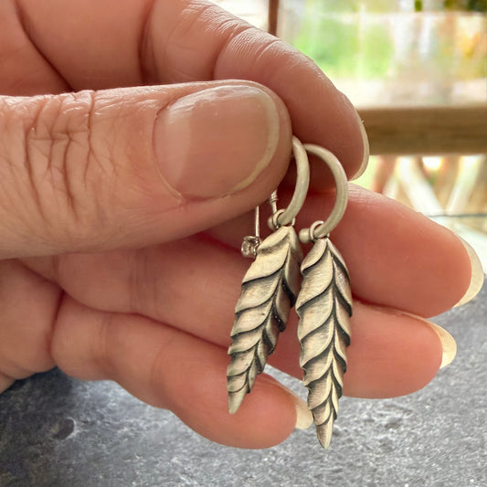 Silver leaf-shaped earrings held between fingers with a blurred background