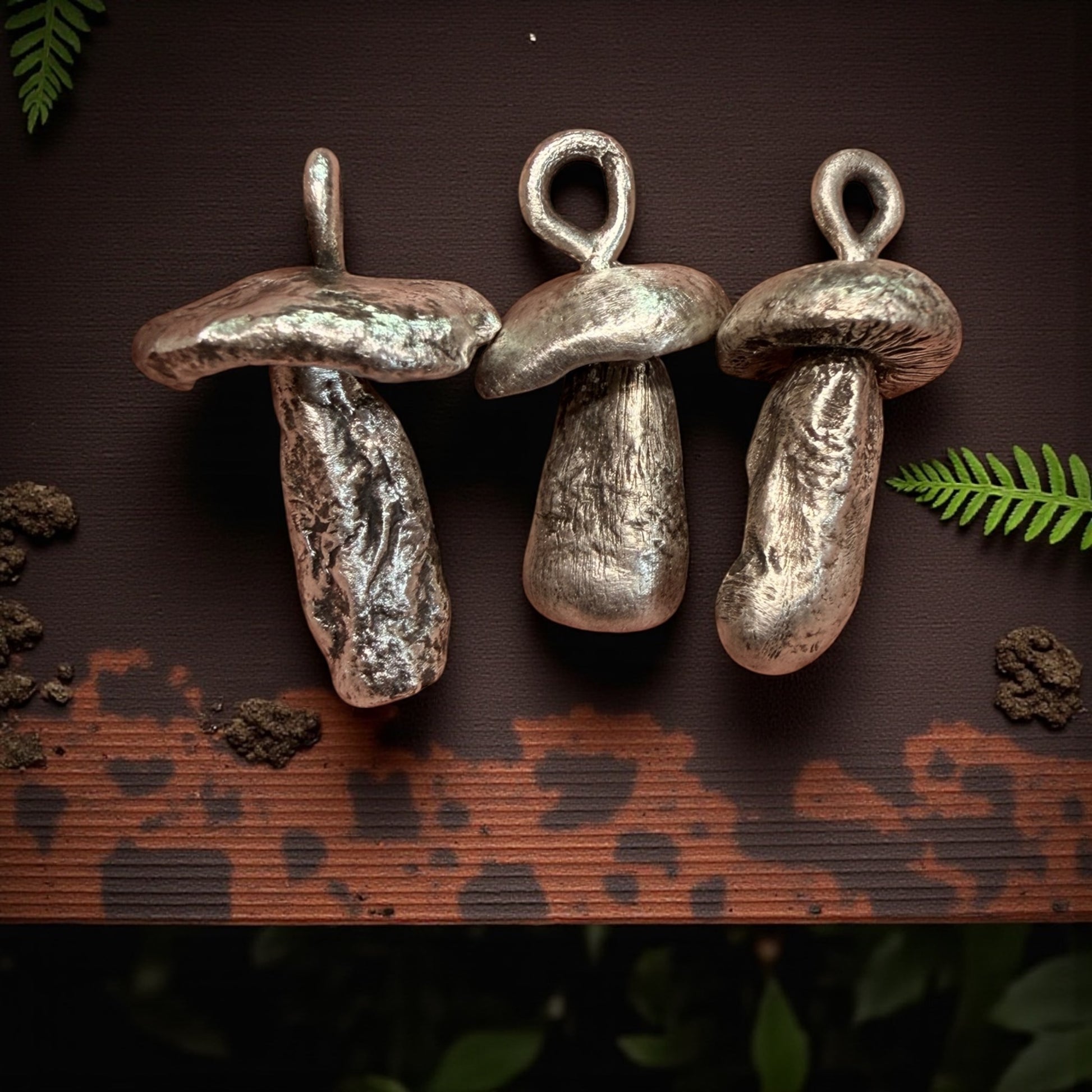 Three silver  mushroom-shaped pendants on a dark brown surface with green leaves.