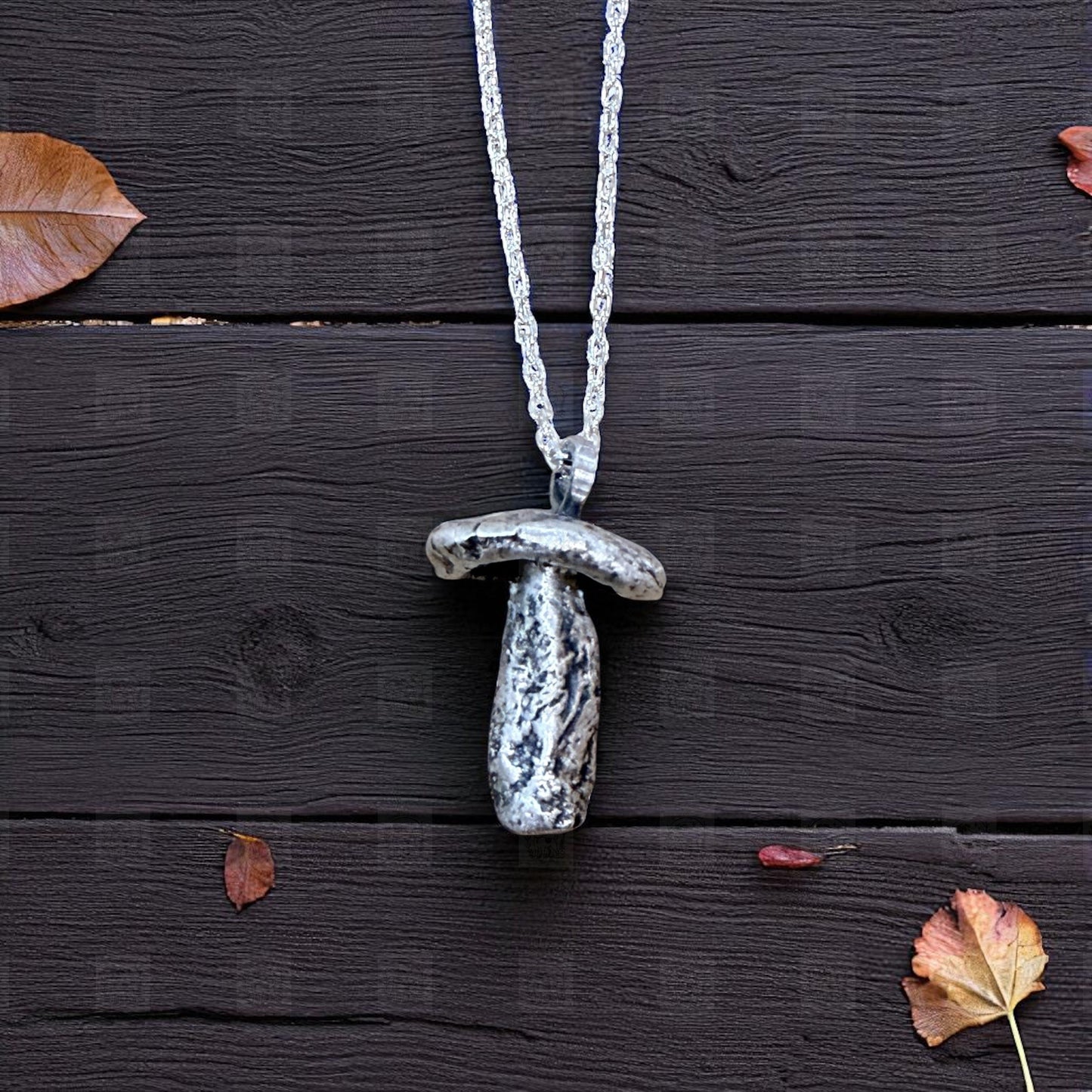 Silver mushroom-shaped pendant necklace on a dark wooden surface with scattered leaves.