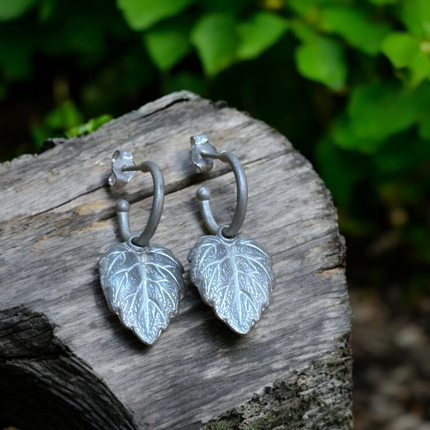 A pair of silver hoop earrings with leaf-shaped charms, placed on a wooden surface with green foliage in the background.