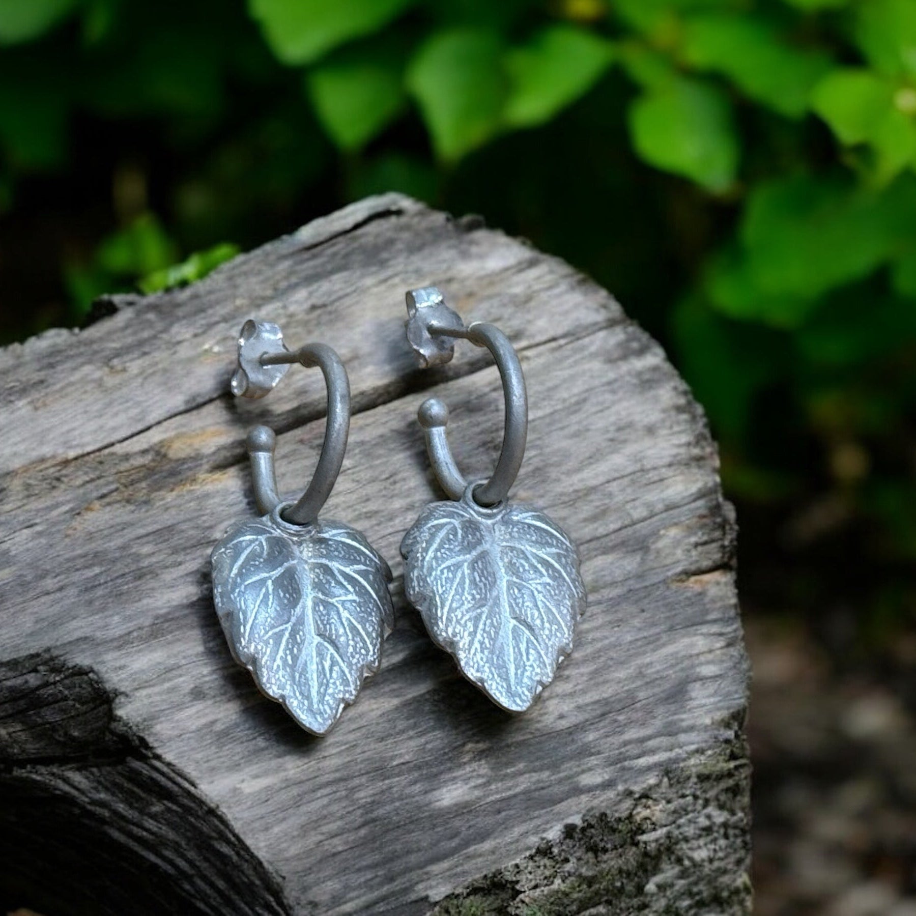 A pair of silver hoop earrings with leaf-shaped charms, placed on a wooden surface with green foliage in the background.