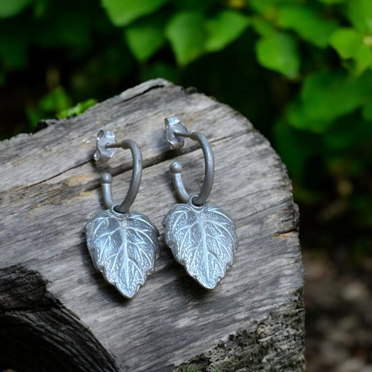 A pair of silver hoop earrings with leaf-shaped charms, placed on a wooden surface with green foliage in the background.