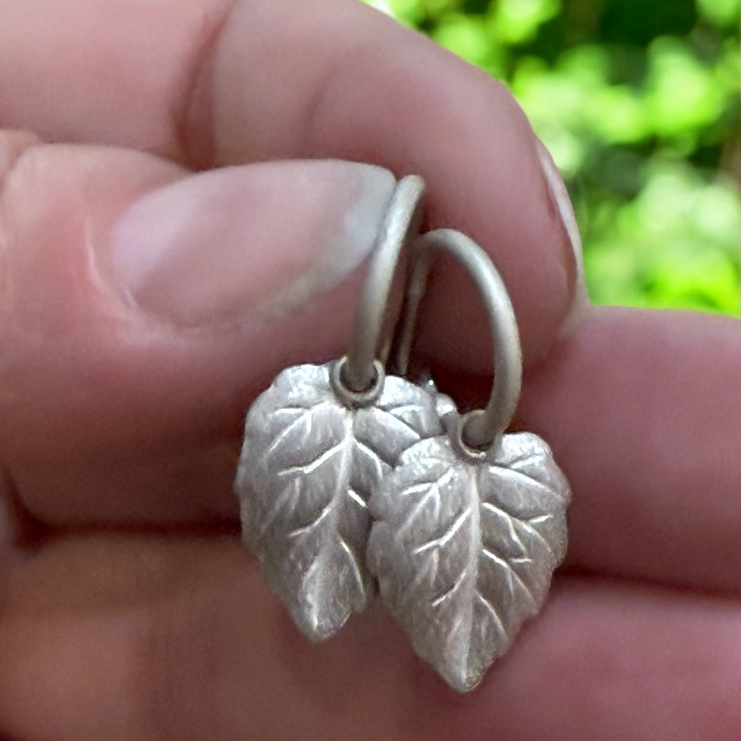 A pair of silver hoop earrings with leaf charms held in front of green foliage.