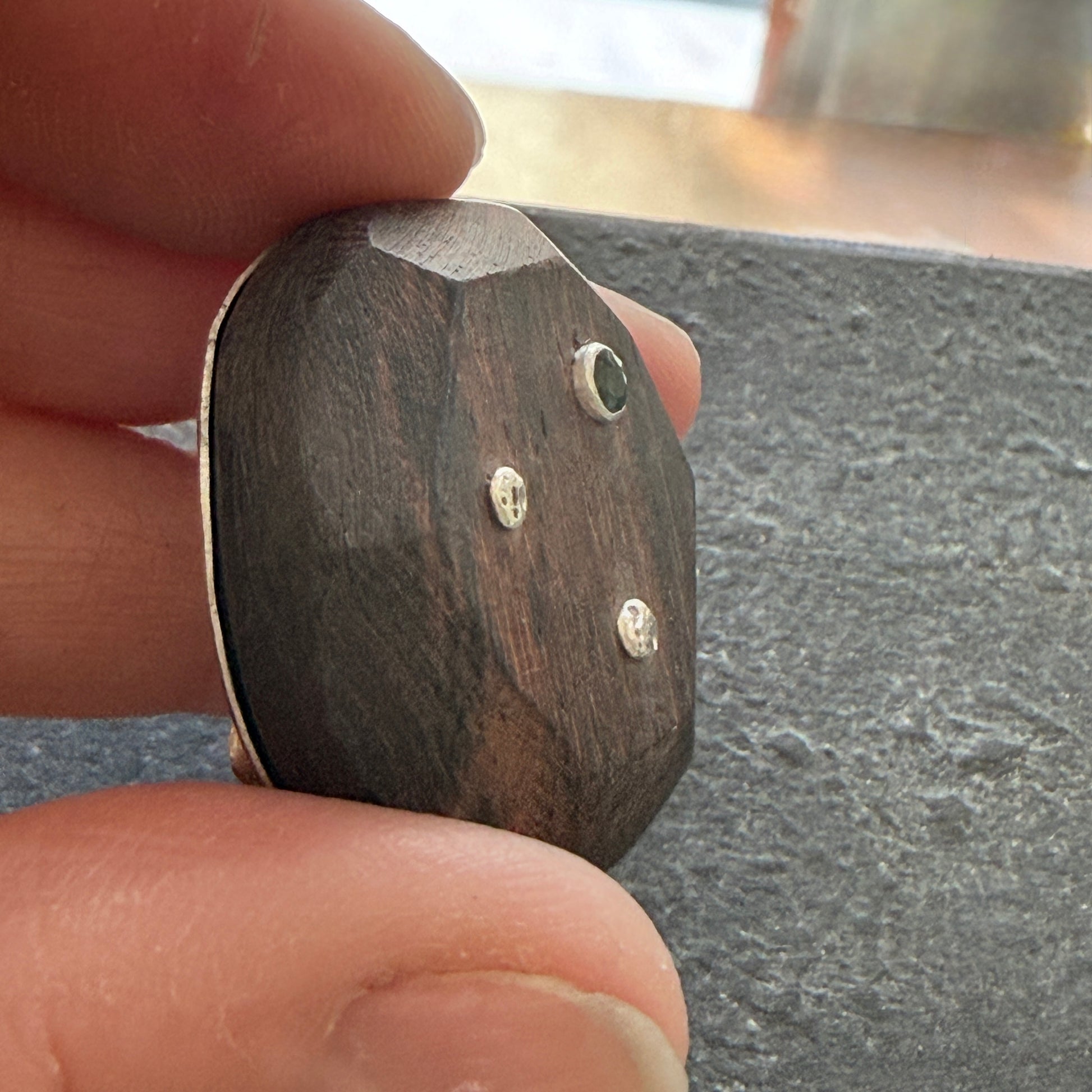 Wooden brooch with sapphire & silver accents held between fingers on a dark surface