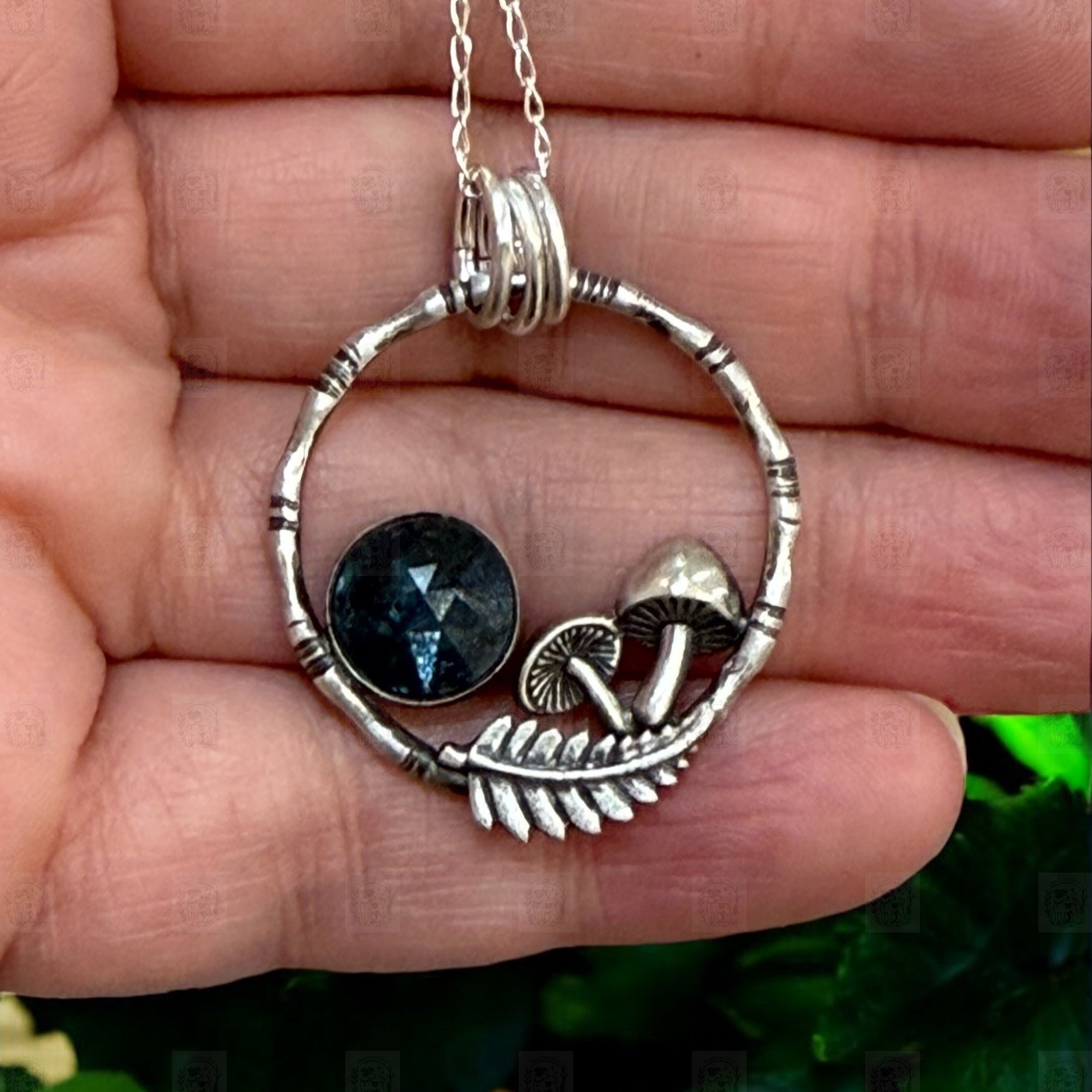 A pendant resting on a hand. The pendant is a bamboo style circle with mushrooms, a fern frond and a teal moss kyanite stone