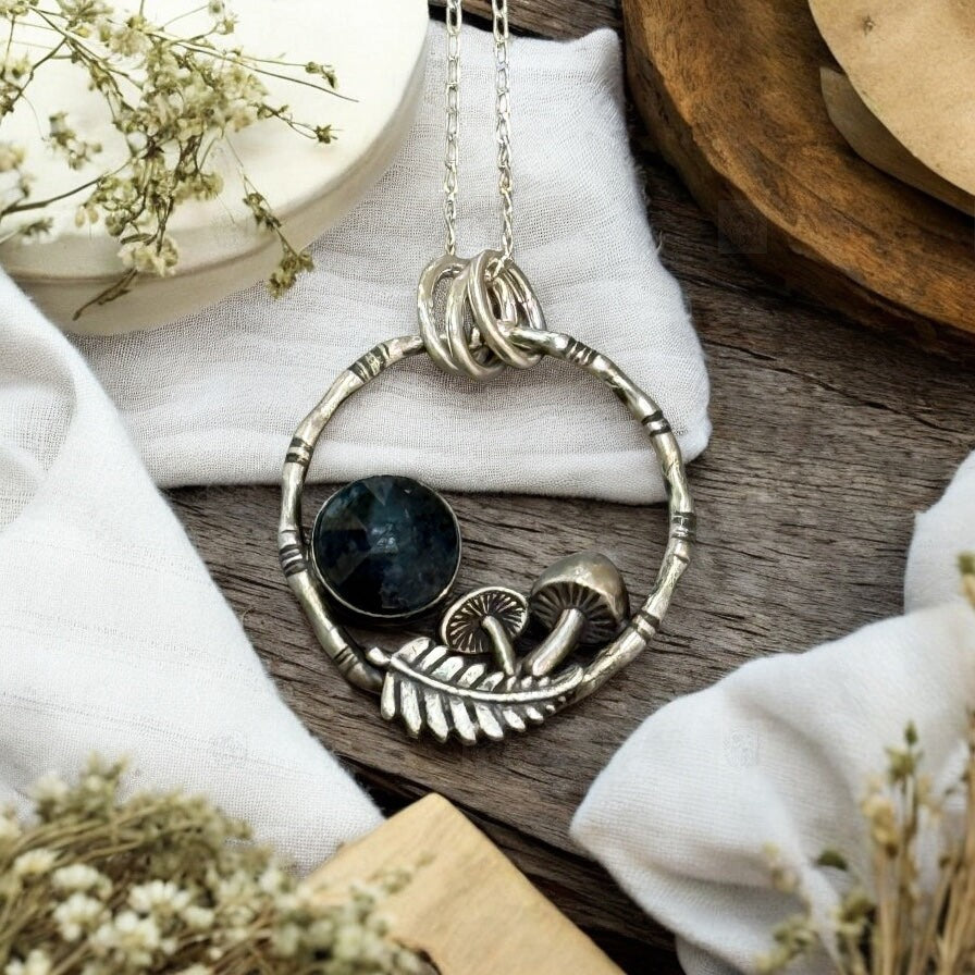 A pendant resting on a table with pale linen and dried flowers. The pendant is a bamboo style circle with mushrooms, a fern frond and a teal moss kyanite stone