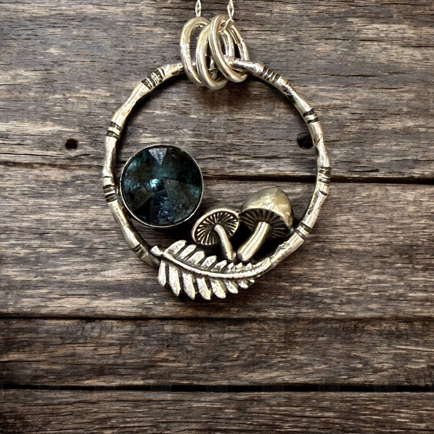 A pendant resting on a dark wooden table. The pendant is a bamboo style circle with mushrooms, a fern frond and a teal moss kyanite stone