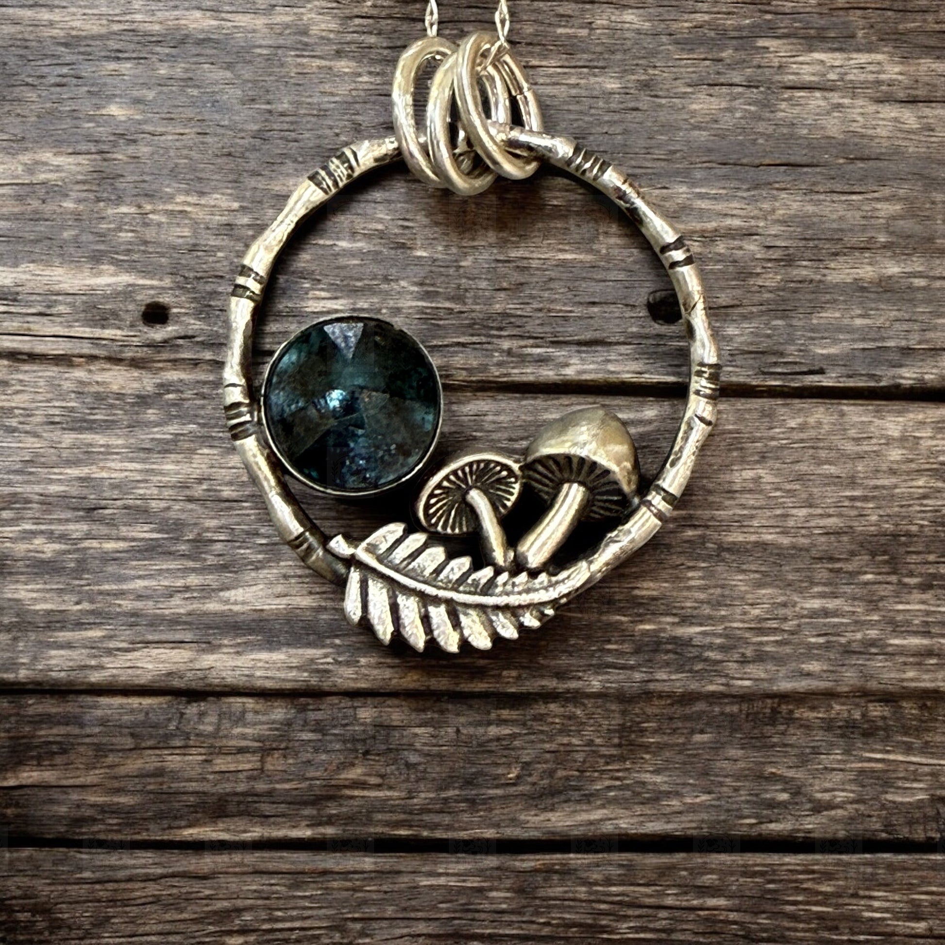 A pendant resting on a dark wooden table. The pendant is a bamboo style circle with mushrooms, a fern frond and a teal moss kyanite stone
