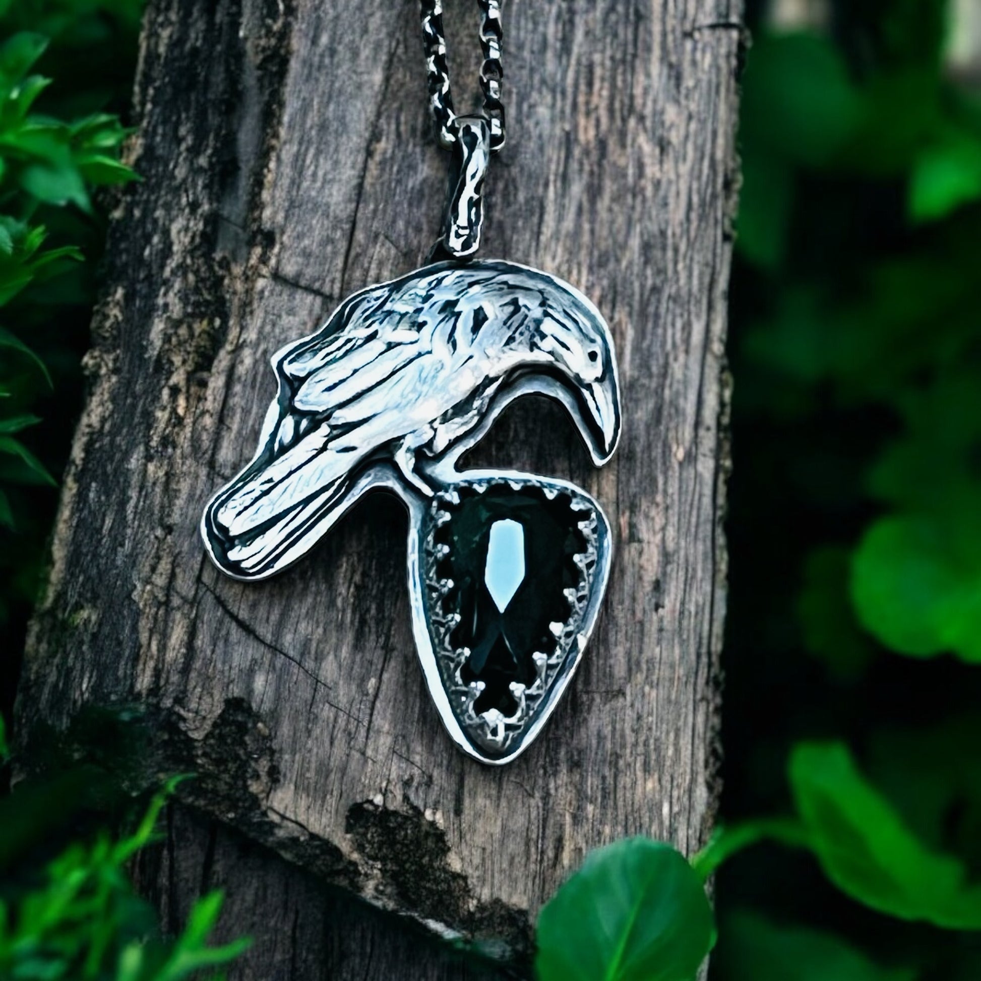 A silver pendant in the shape of a raven perched on a piece of wood, featuring a large green tourmaline stone in the center.