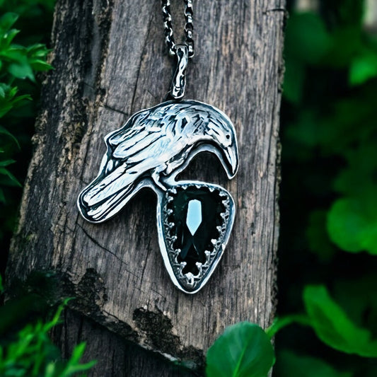 A silver pendant in the shape of a raven perched on a piece of wood, featuring a large green tourmaline stone in the center.