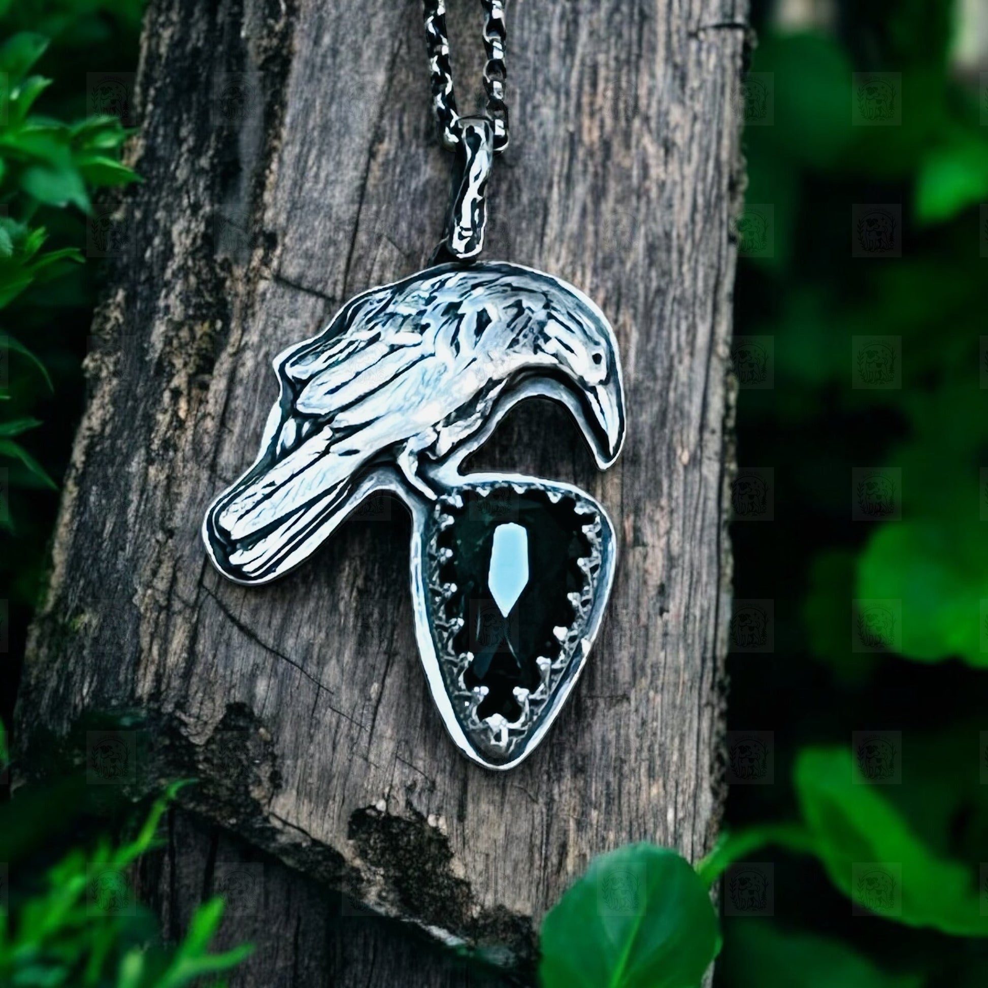 A silver pendant in the shape of a raven perched on a piece of wood, featuring a large green tourmaline stone in the center.