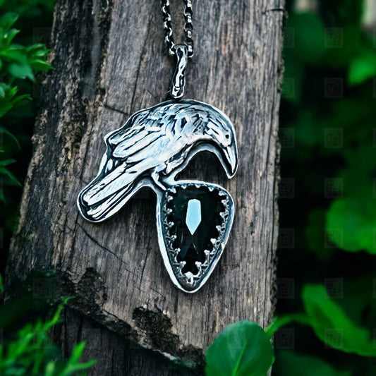 A silver pendant in the shape of a raven perched on a piece of wood, featuring a large green tourmaline stone in the center.
