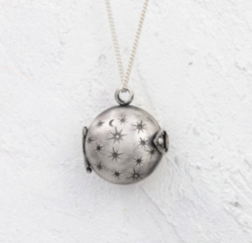 A domed silver locket with a star & moon design is suspended in front of a white wall.