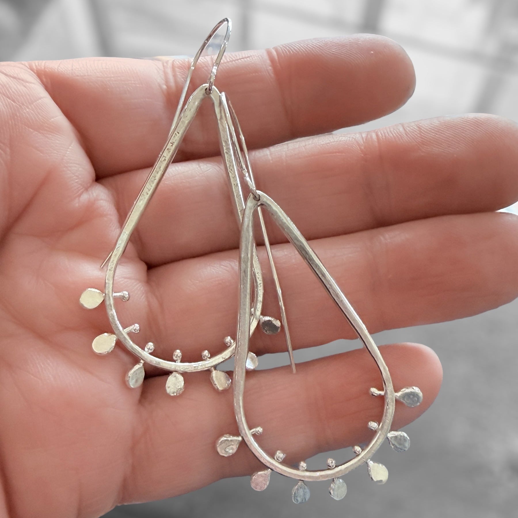 Silver teardrop earrings held in a hand against a blurred background.