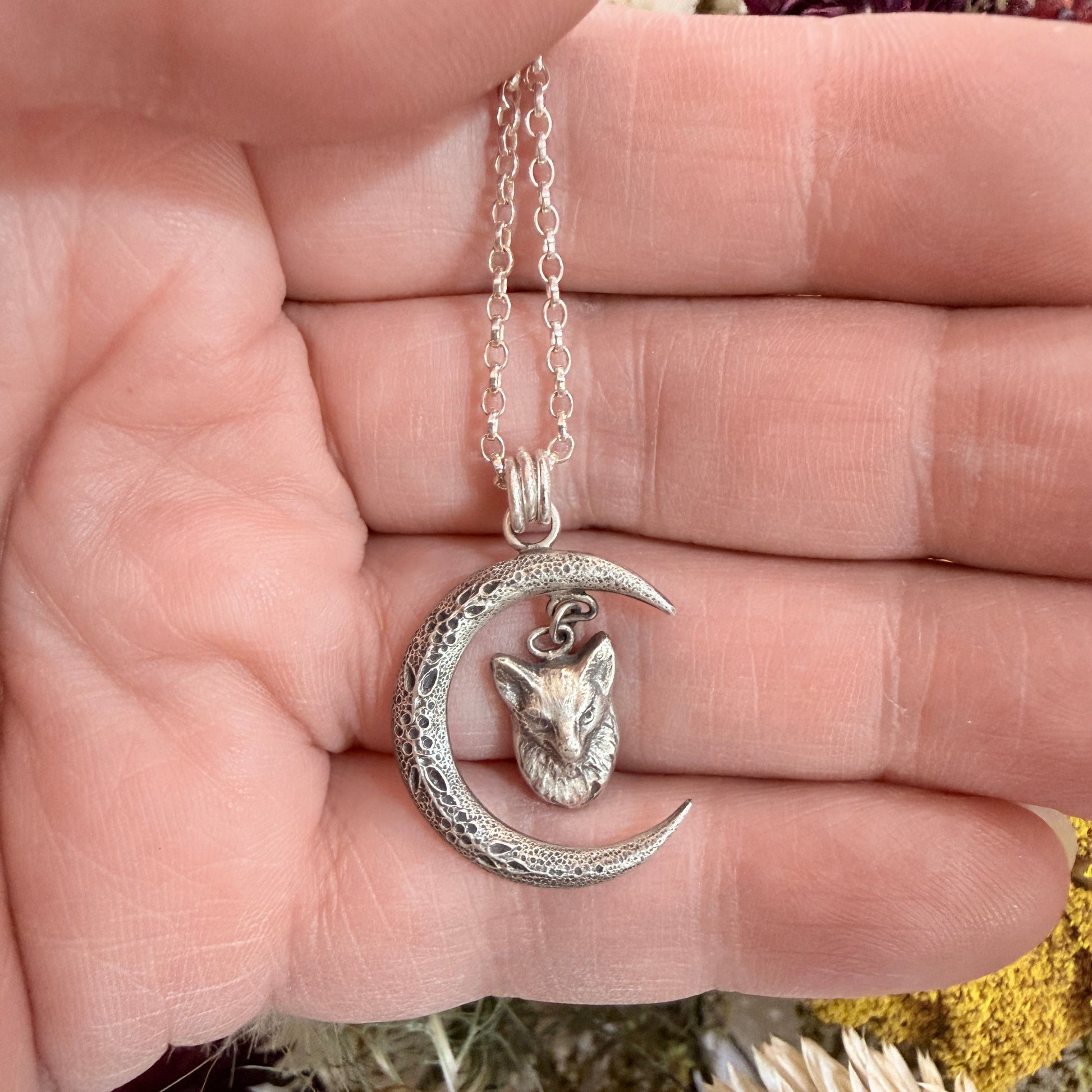 Silver necklace with a fox and moon pendant held in a hand, with dried flowers in the background.