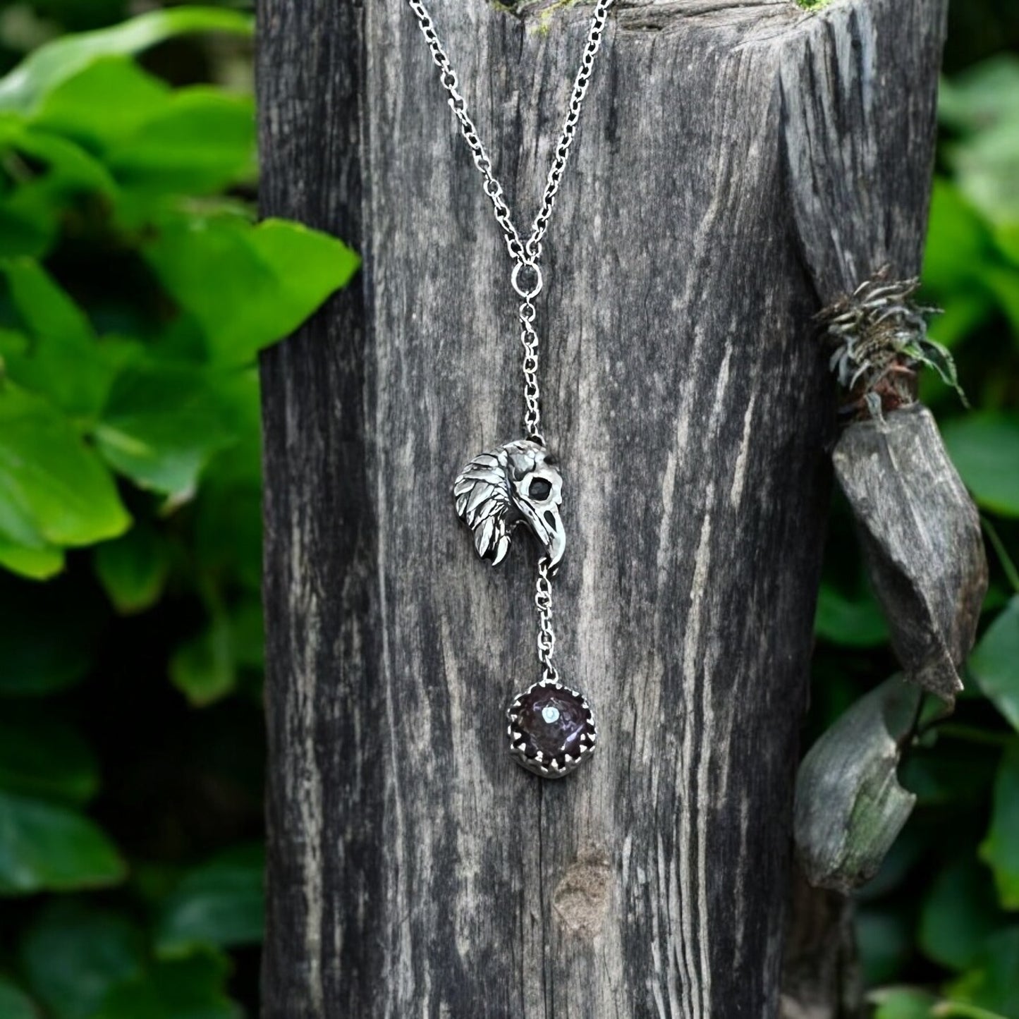 A sterling silver raven's head pendant with a purple red ruby attached to a silver chain, displayed on a wooden post with green foliage in the background.