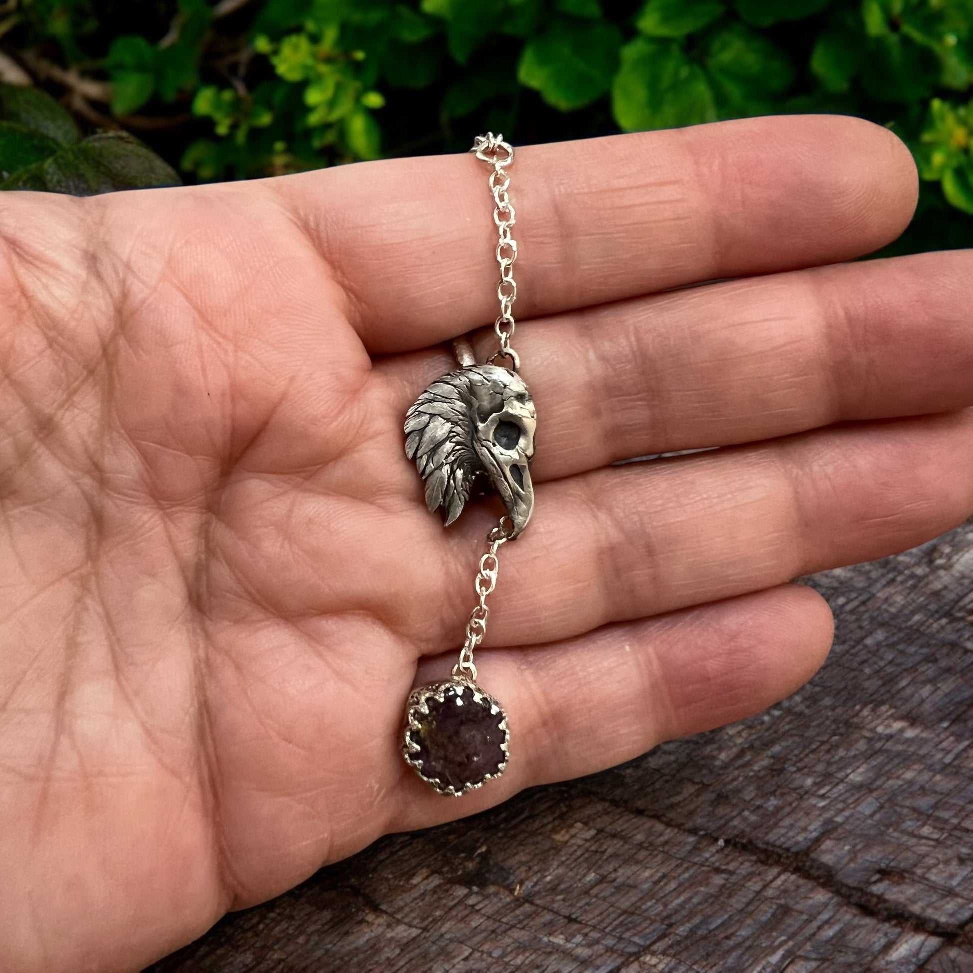 A raven head pendant held in front of wood & foliage. 