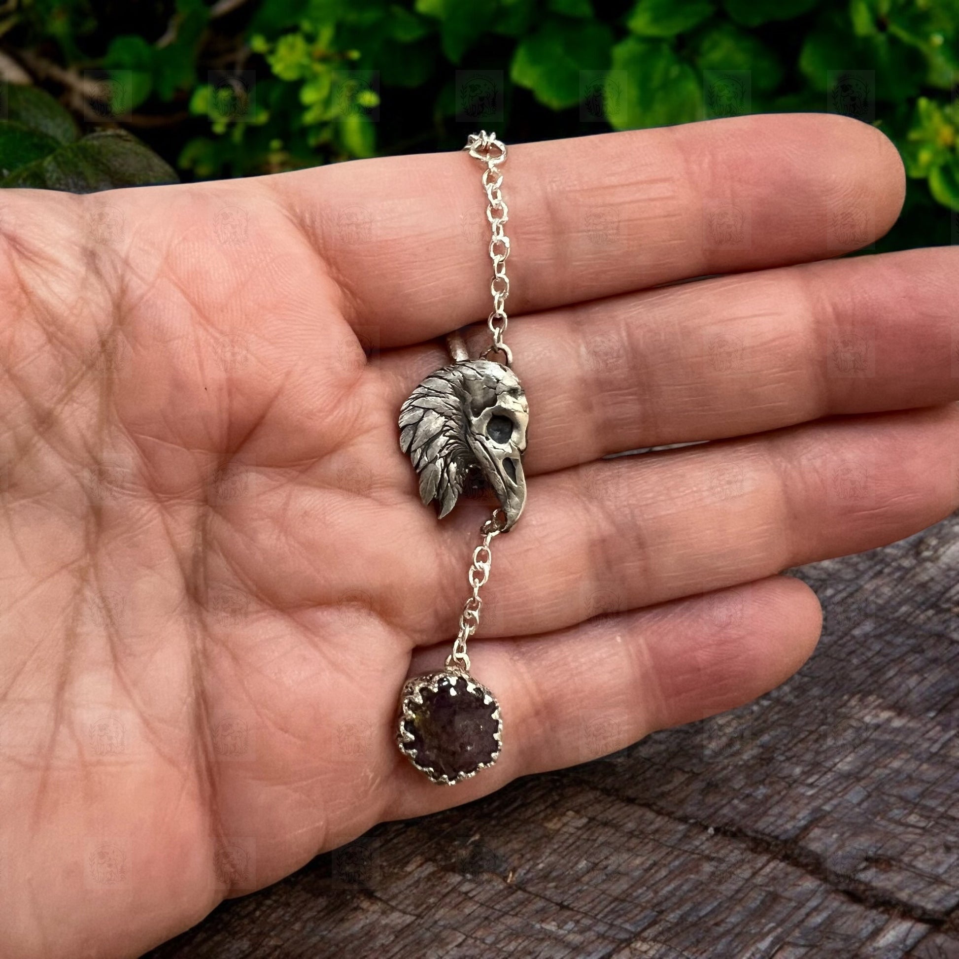 A raven head pendant held in front of wood & foliage.