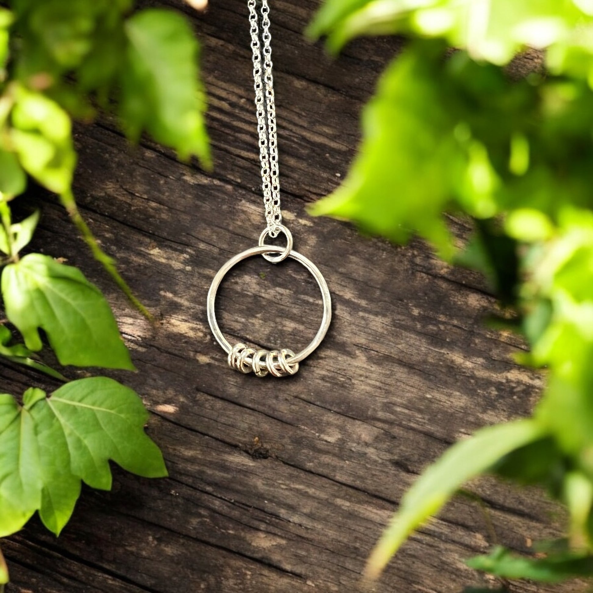 a sterling silver necklace with 5 silver rings sits on a distressed wooden surface surrounded with greenery.