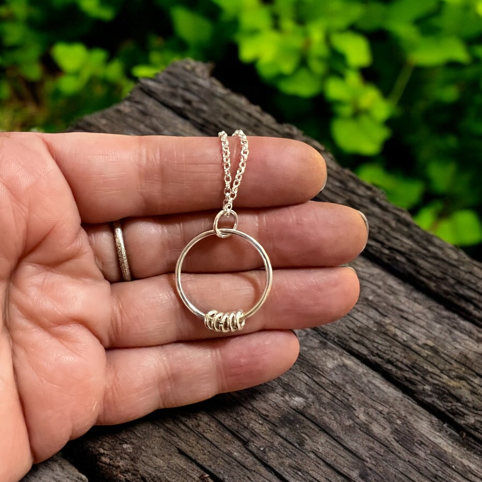 A sterling silver necklace held in front of a distressed wooden surface with greenery behind.