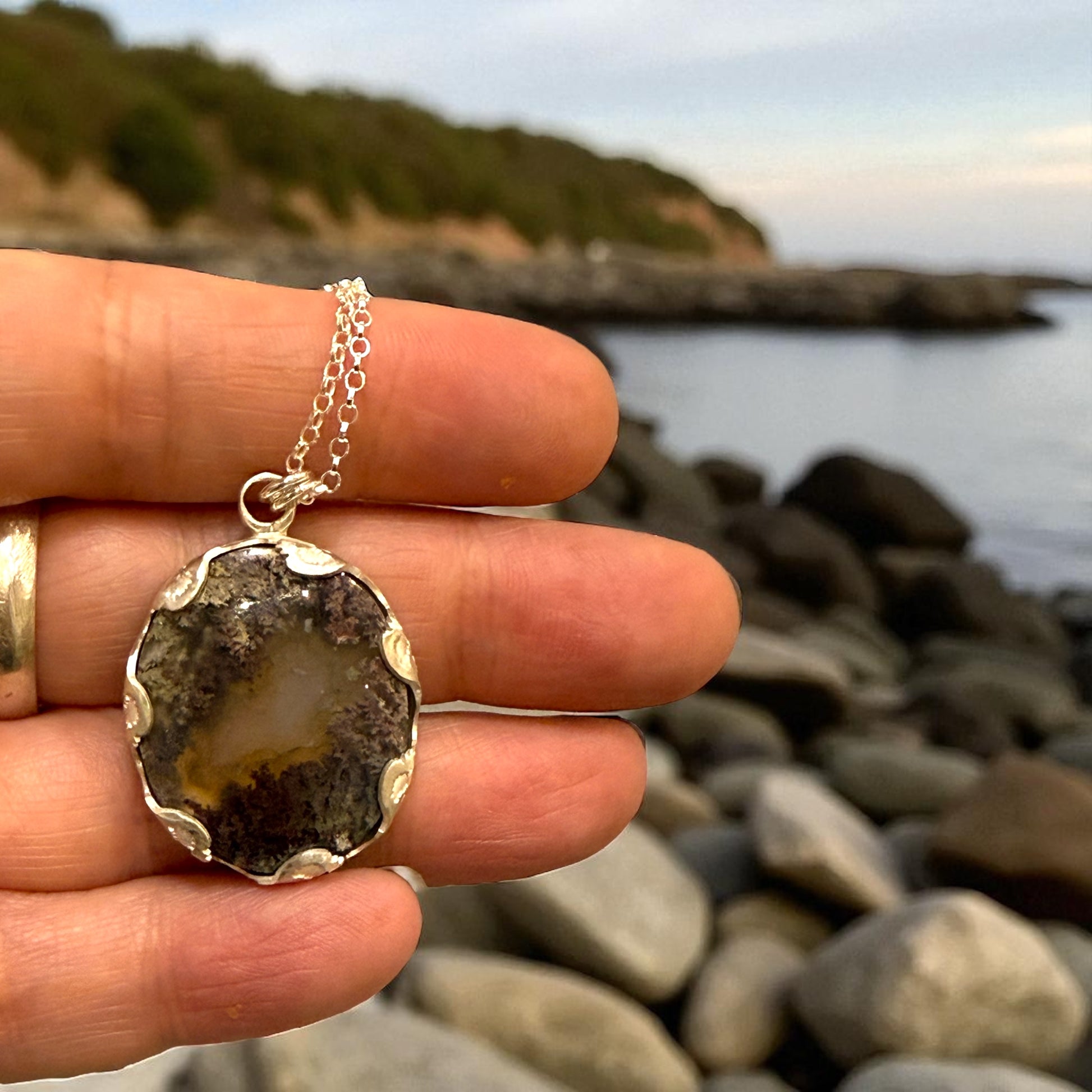 Hand holding a silver pendant with a natural stone design against a rocky beach background