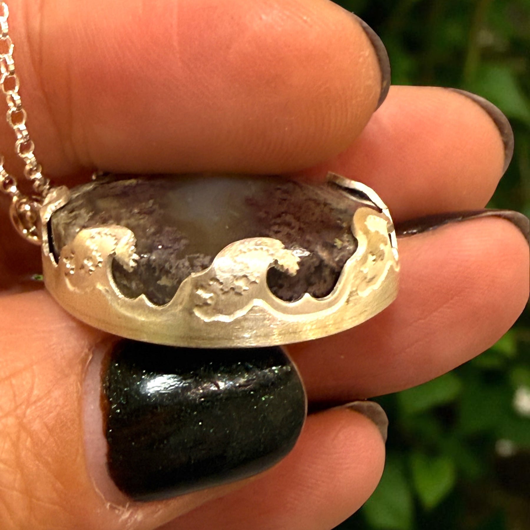 Silver necklace with a pendant held between fingers against a blurred green background