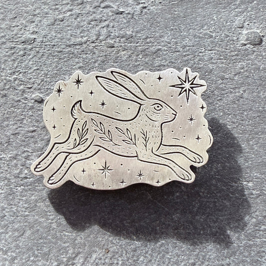 Sterling silver hare brooch with star and leaf design on a textured gray background